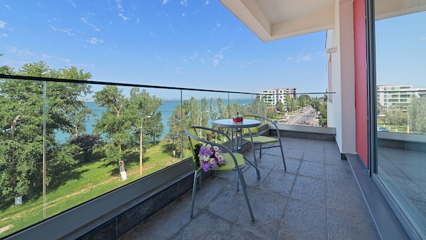 A balcony with a glass railing, a table, and chairs. There is a view of a body of water and greenery. - Tomis Garden Apartments (Constanta)
