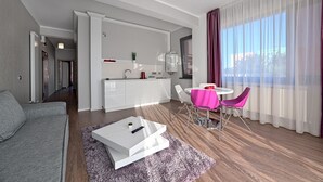 A modern hotel room with a sofa, a dining table with chairs, a kitchen area, and a large window with curtains. - Tomis Garden Apartments (Constanta)