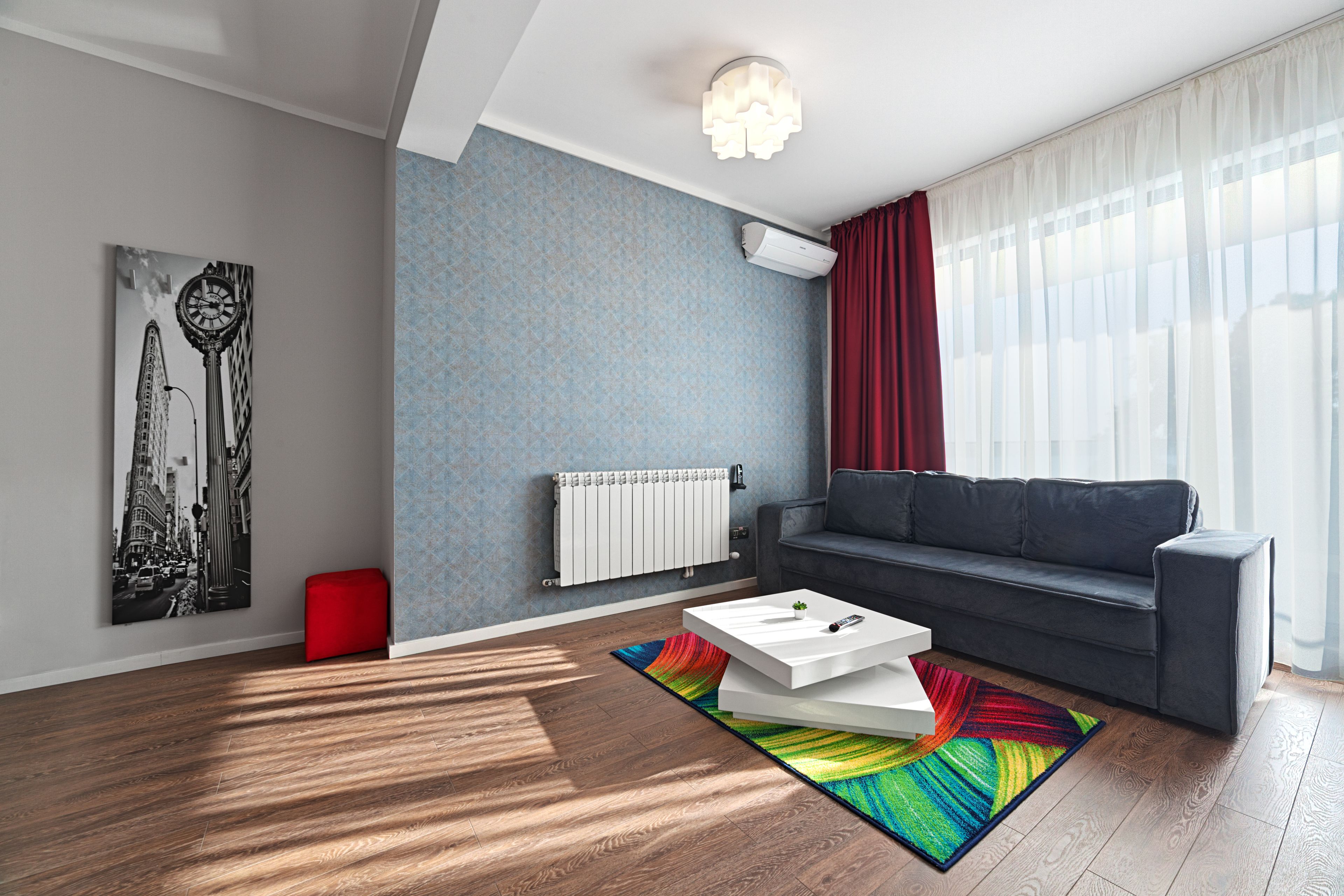 A modern living room with a grey sofa, a white coffee table, a colorful rug, a red ottoman, and a cityscape painting on the wall.