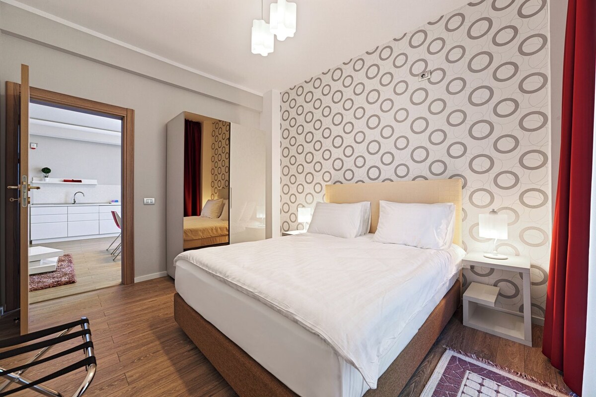 Un hotel moderno con una cama grande, una mesita de noche con lámpara, una cortina roja y un papel tapiz estampado.