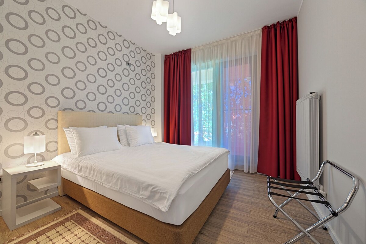 Une chambre d’hôtel avec un grand lit, une table de chevet avec une lampe, un radiateur et une fenêtre avec des rideaux rouges.