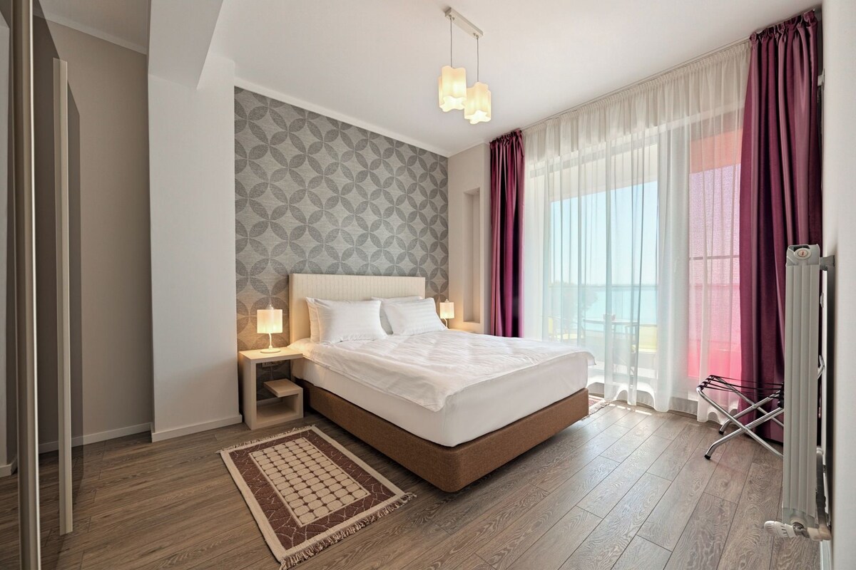 Habitación de hotel moderna con una cama grande, una mesita de noche, un velador y un papel tapiz decorativo, con vistas al mar a través de cortinas translúcidas.