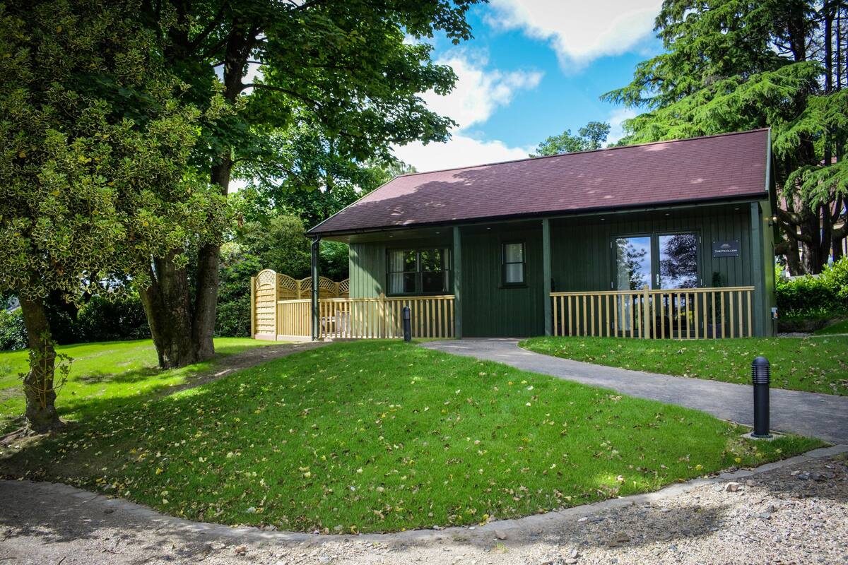 A small building with a green exterior and a dark roof, surrounded by trees and a grassy area.