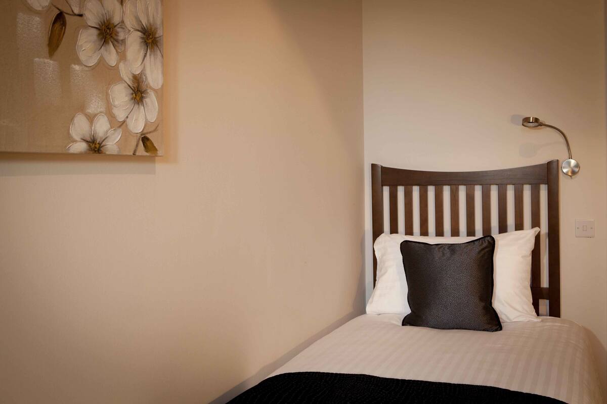 A neatly made bed with a striped headboard, a wall-mounted lamp, and a floral painting above it.