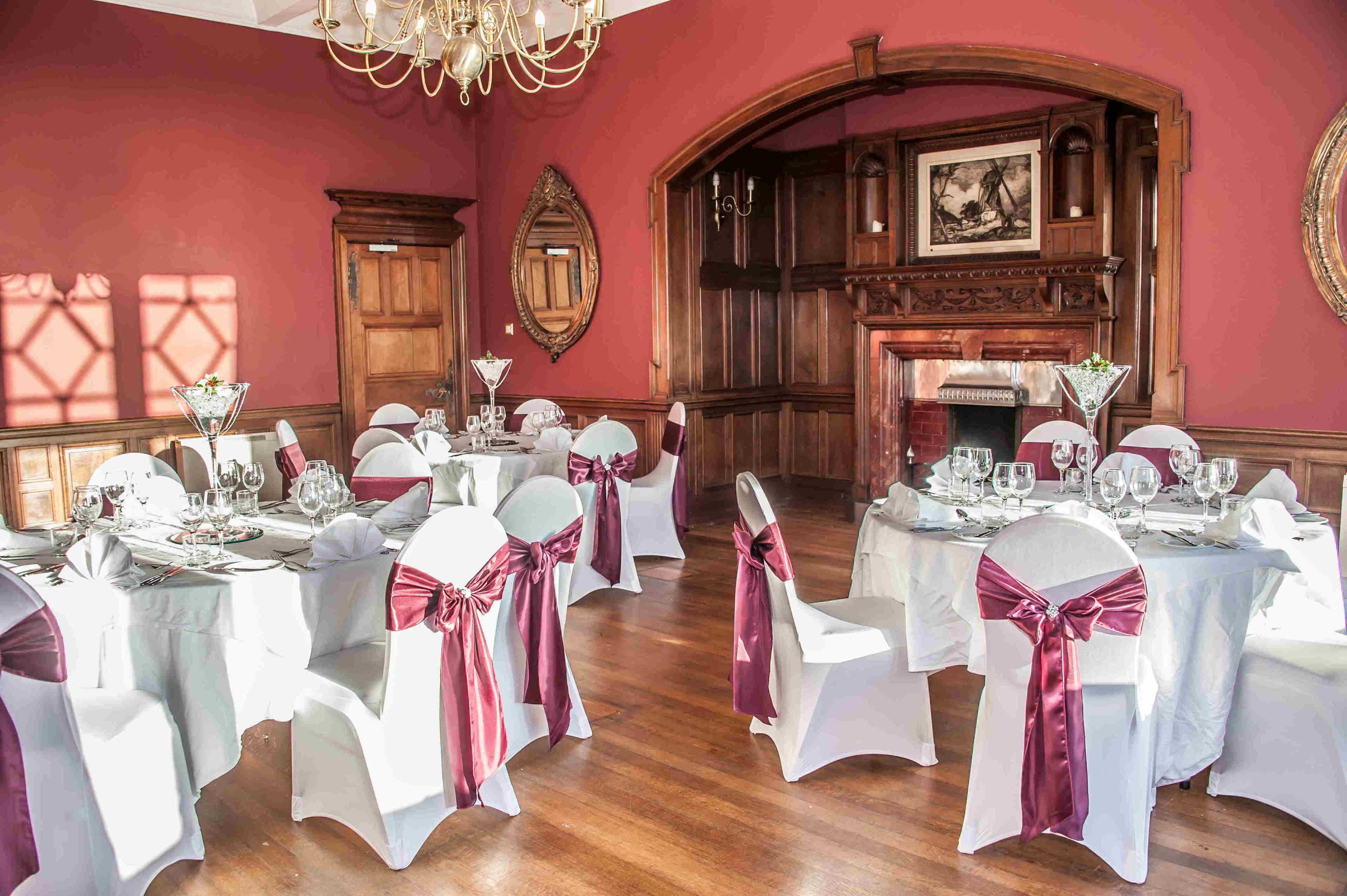 Una sala da pranzo formale con diversi tavoli apparecchiati per un pasto, sedie bianche con nastri rossi e un camino in legno.