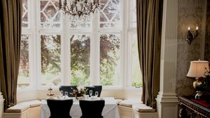 A dining room with a table set for two, a chandelier, and a view of trees through large windows.