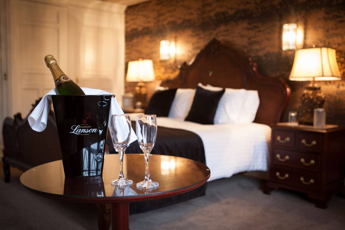A hotel room with a bed, bedside lamps, a round table with a champagne bottle and two glasses, and a nightstand with a lamp.