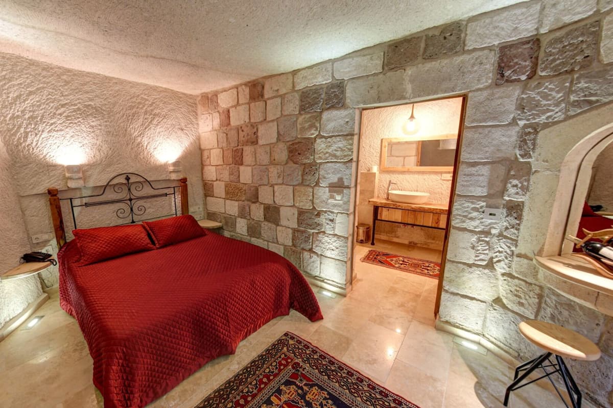 Une chambre avec un mur en pierre, un lit recouvert d’une couverture rouge, une table de chevet, un tabouret en bois et une salle de bain visible à travers une arche.