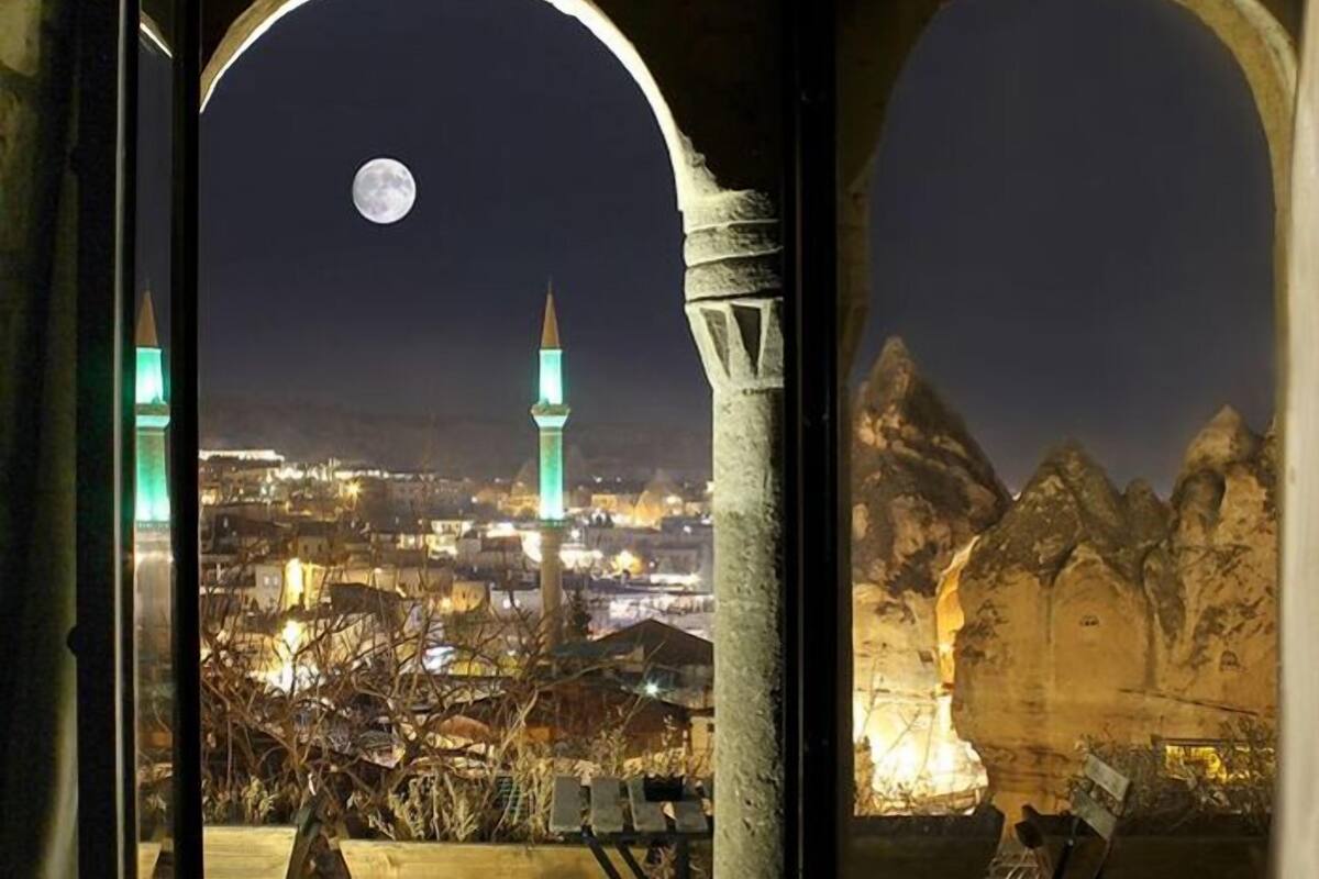 Une vue nocturne depuis un balcon, avec la lune et le paysage urbain en arrière-plan, comprenant une mosquée et des montagnes.