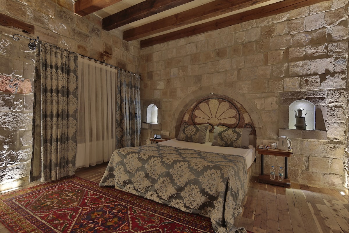Une chambre avec un mur en pierre, un cadre de lit en bois, une couvre-lit à motifs, un tapis rouge et beige, et une petite table de chevet avec une lampe.