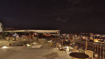 An outdoor seating area with tables and chairs, a covered section, and a view of a town with buildings and a mountain in the background.