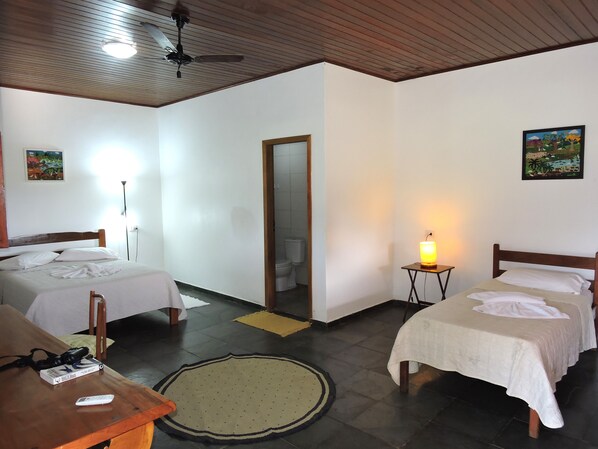 A hotel room with two beds, a wooden ceiling, a ceiling fan, a small table with a remote control, and a painting on the wall.