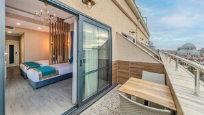 A balcony with a wooden table and chairs, a glass door leading to a bedroom with a bed and a chandelier, and a cityscape view in the background. - Marnas Hotels (Istanbul)