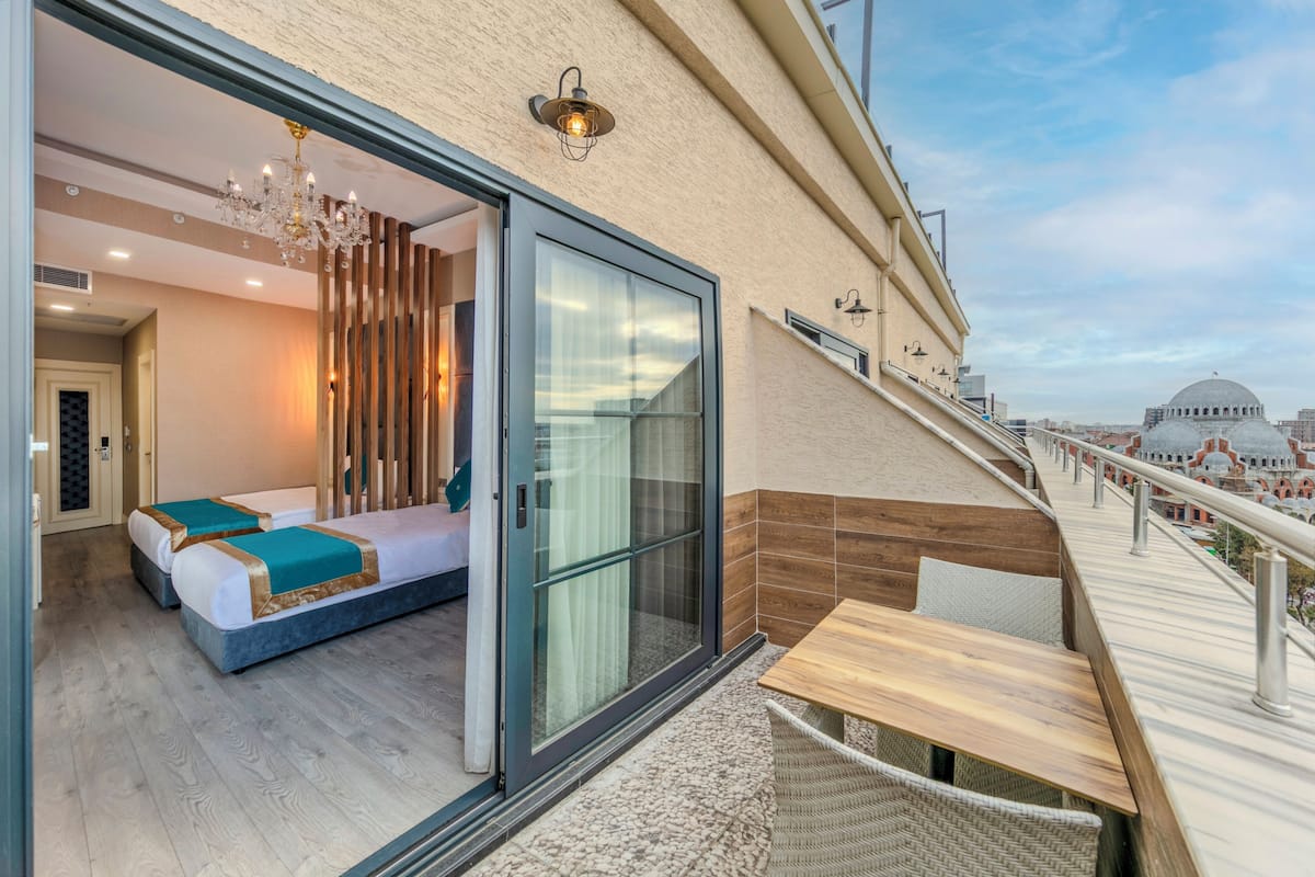 A balcony with a wooden table and chairs, a glass door leading to a bedroom with a bed and a chandelier, and a cityscape view in the background.