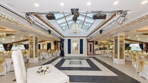 A spacious banquet hall with a high ceiling, chandeliers, and a central decorative wall. Tables are set with white tablecloths and gold sashes. - Marnas Hotels (Istanbul)