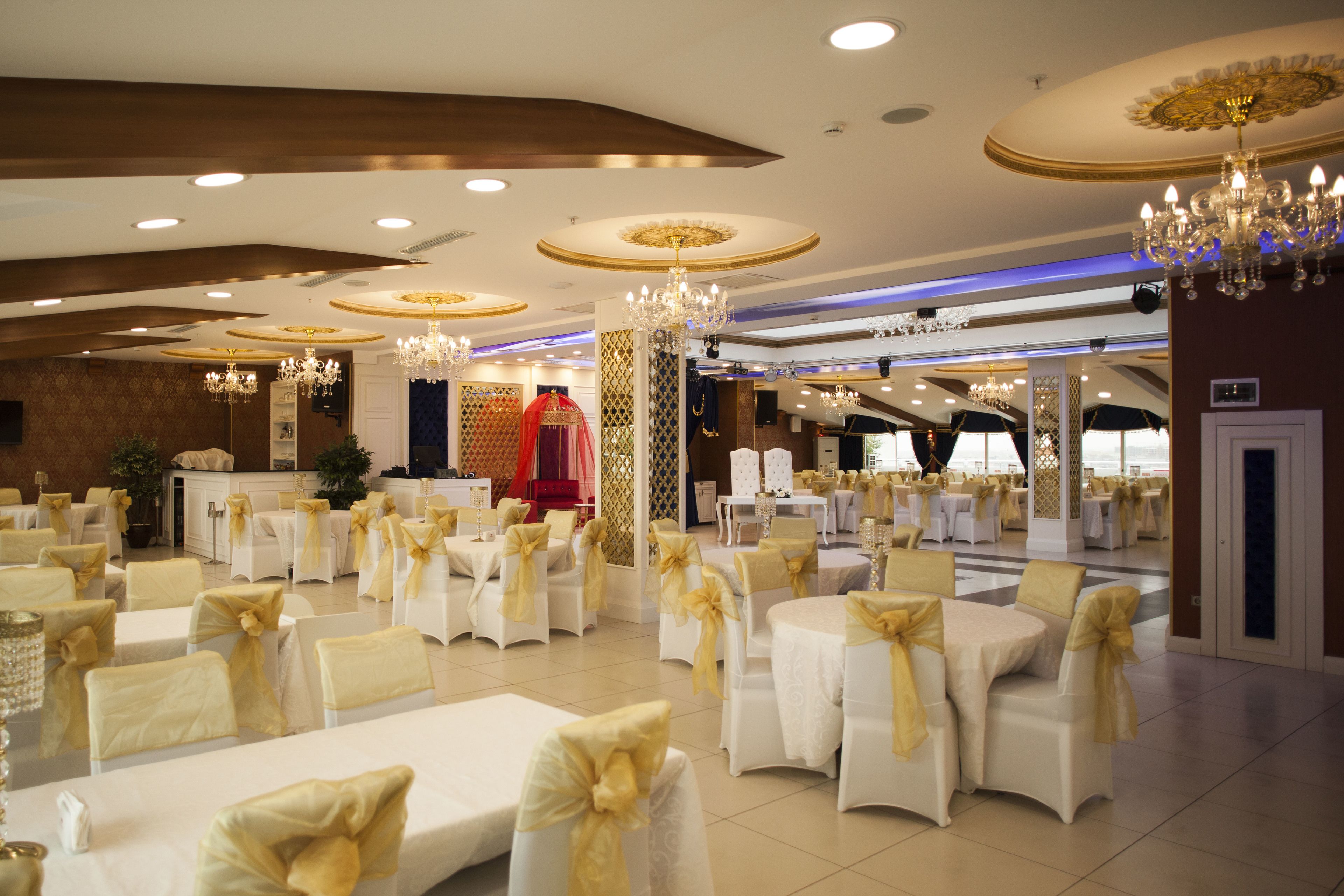 A banquet hall with round tables, white chairs with gold sashes, chandeliers, and a red-clad figure in the background.