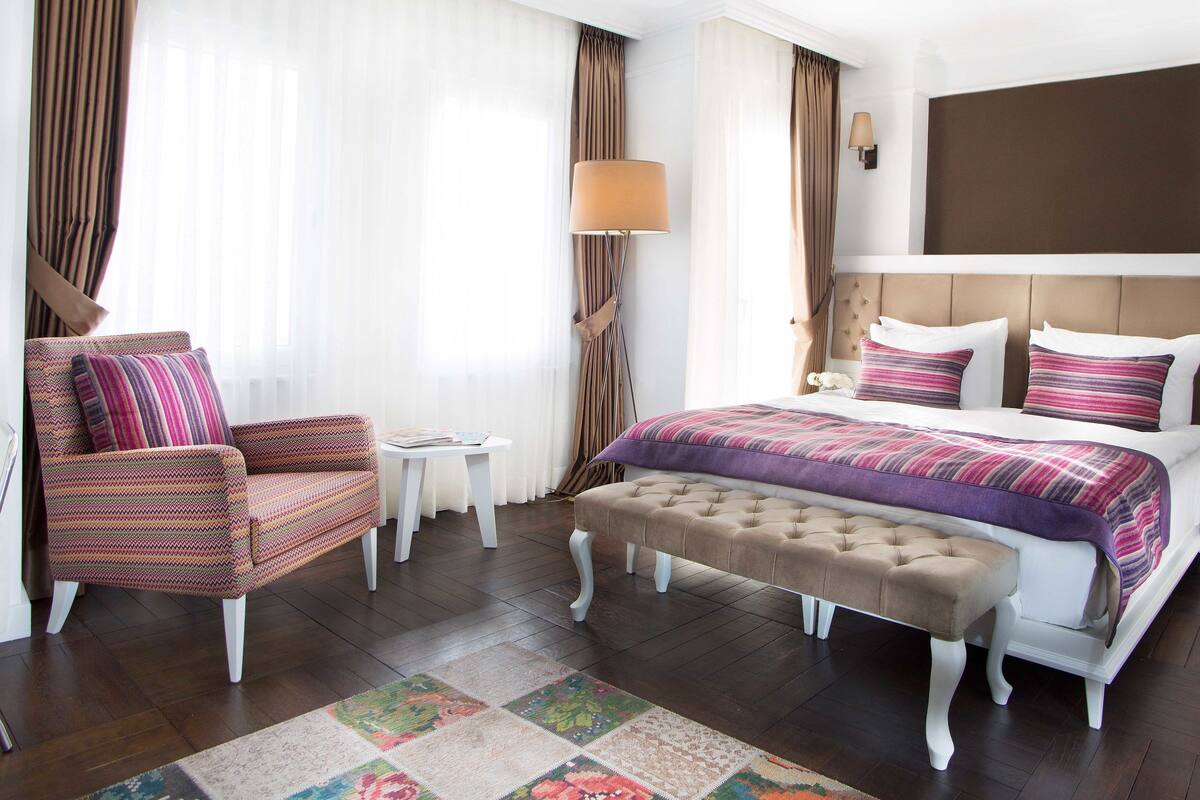 A hotel room with a bed, a chair, a nightstand, a lamp, and a large window with curtains.