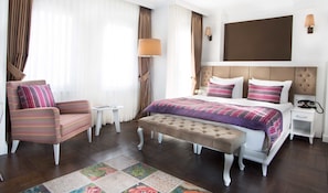 Quarto de hotel com uma cama, uma cadeira, uma mesa de cabeceira, um abajur e uma janela grande com cortinas.