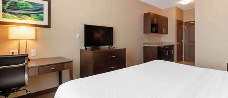 A hotel room with a bed, desk, chair, TV, kitchenette, and a painting on the wall.