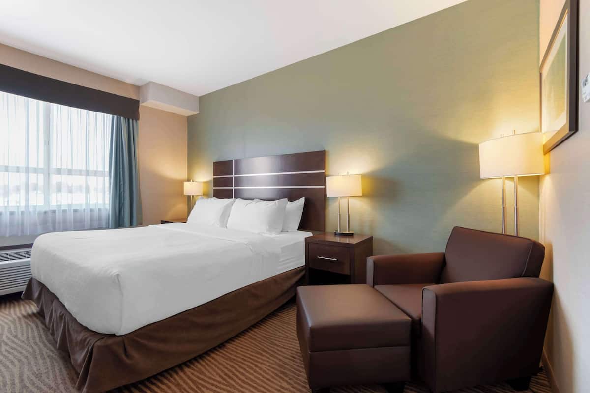 Habitación de hotel con una cama grande, dos mesitas de noche con lámparas, un sillón marrón y vistas al exterior a través de una ventana con cortinas traslúcidas.
