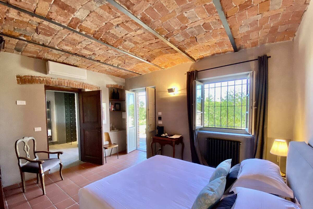 A bedroom with a brick ceiling, a large bed with pillows, a chair, a desk, and a window with a view of greenery.