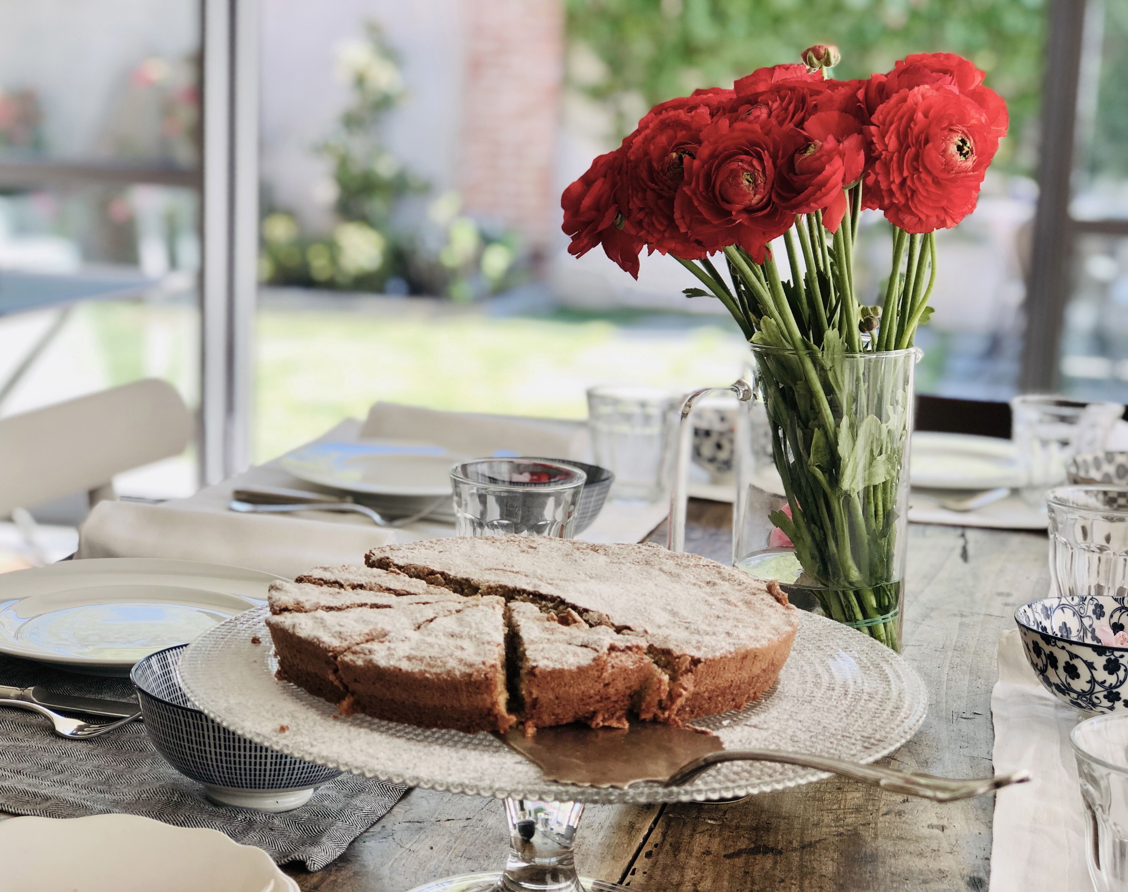 Una mesa con una porción de pastel sobre un plato de vidrio, un jarrón con flores rojas y una mesa de madera rústica.