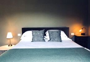Een net opgemaakt bed met een witte deken en twee grijze sierkussens met bloemenprint, een blauwgroene plaid en een bedlampje op een nachtkastje.