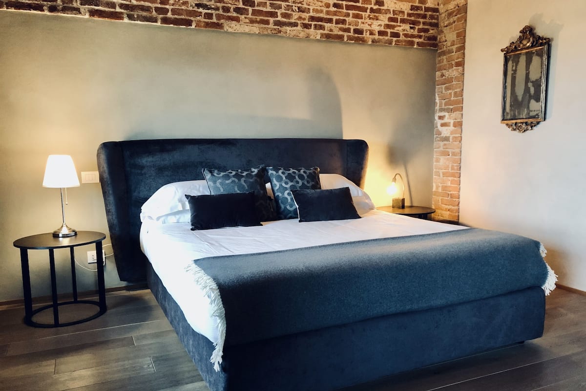 A large bed with a dark headboard, white linens, and multiple pillows. There is a small round table with a lamp beside the bed. The room has a brick wall and a framed picture on the wall.