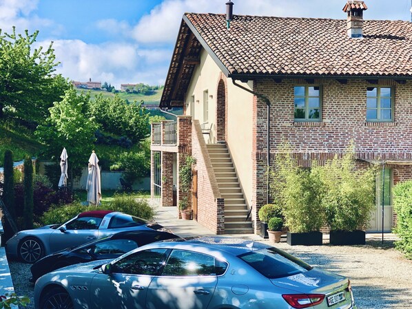 Casa de dos pisos con fachada de ladrillo, techo de tejas y una escalera visible que lleva al piso superior. Hay dos coches estacionados al frente, y la propiedad está rodeada de vegetación y un cielo despejado.