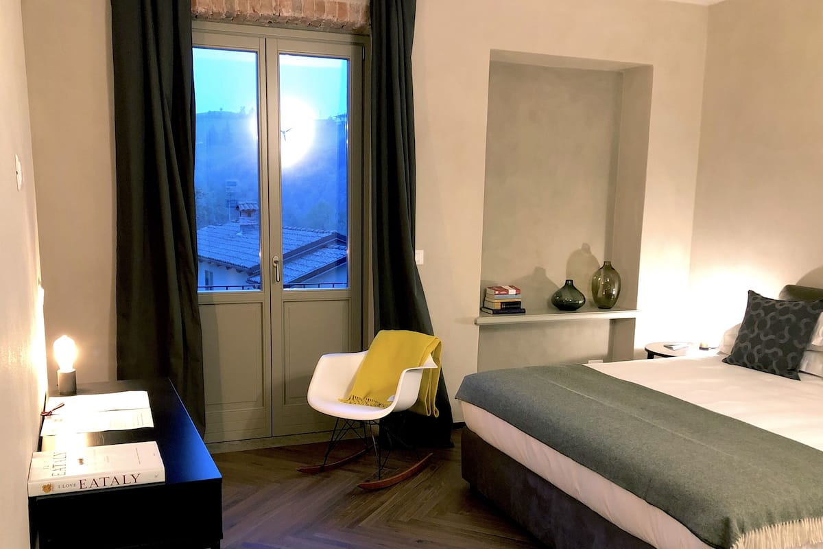A hotel room with a bed, a rocking chair, a desk with books, a window with curtains, and a nightstand with a lamp.