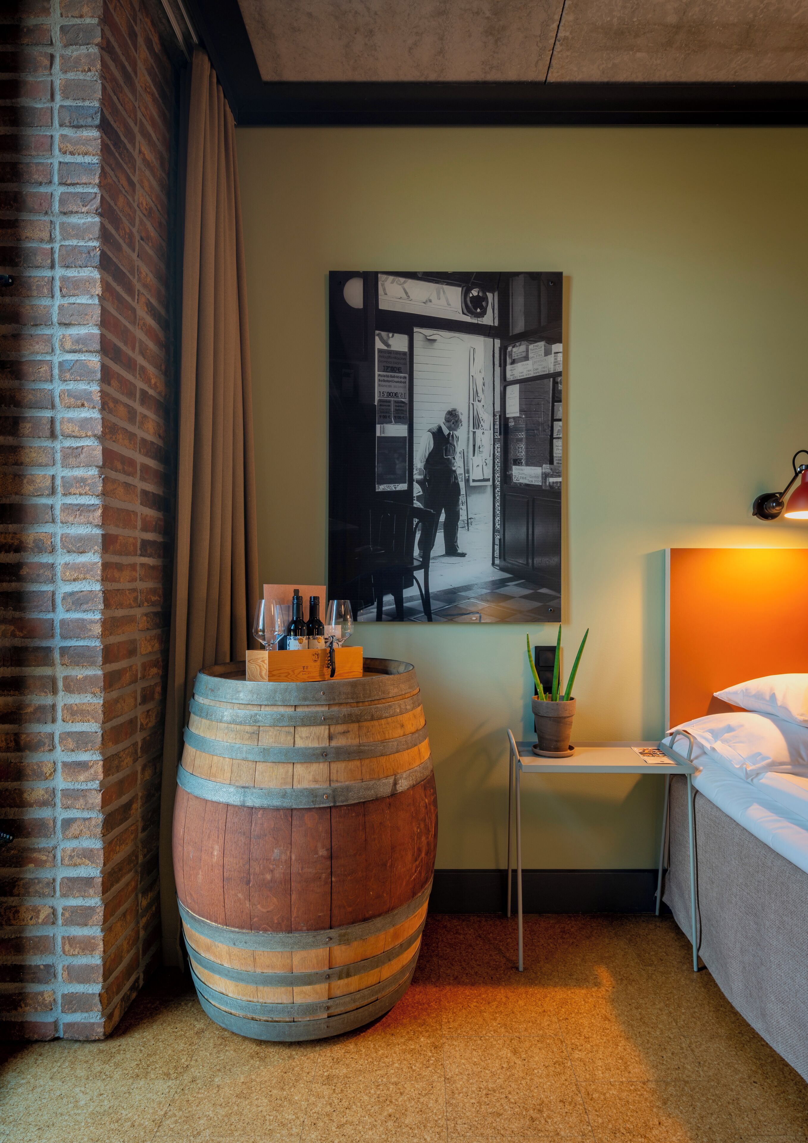 A room with a large wooden barrel, a bed, a small table with a plant, and a wall-mounted picture.