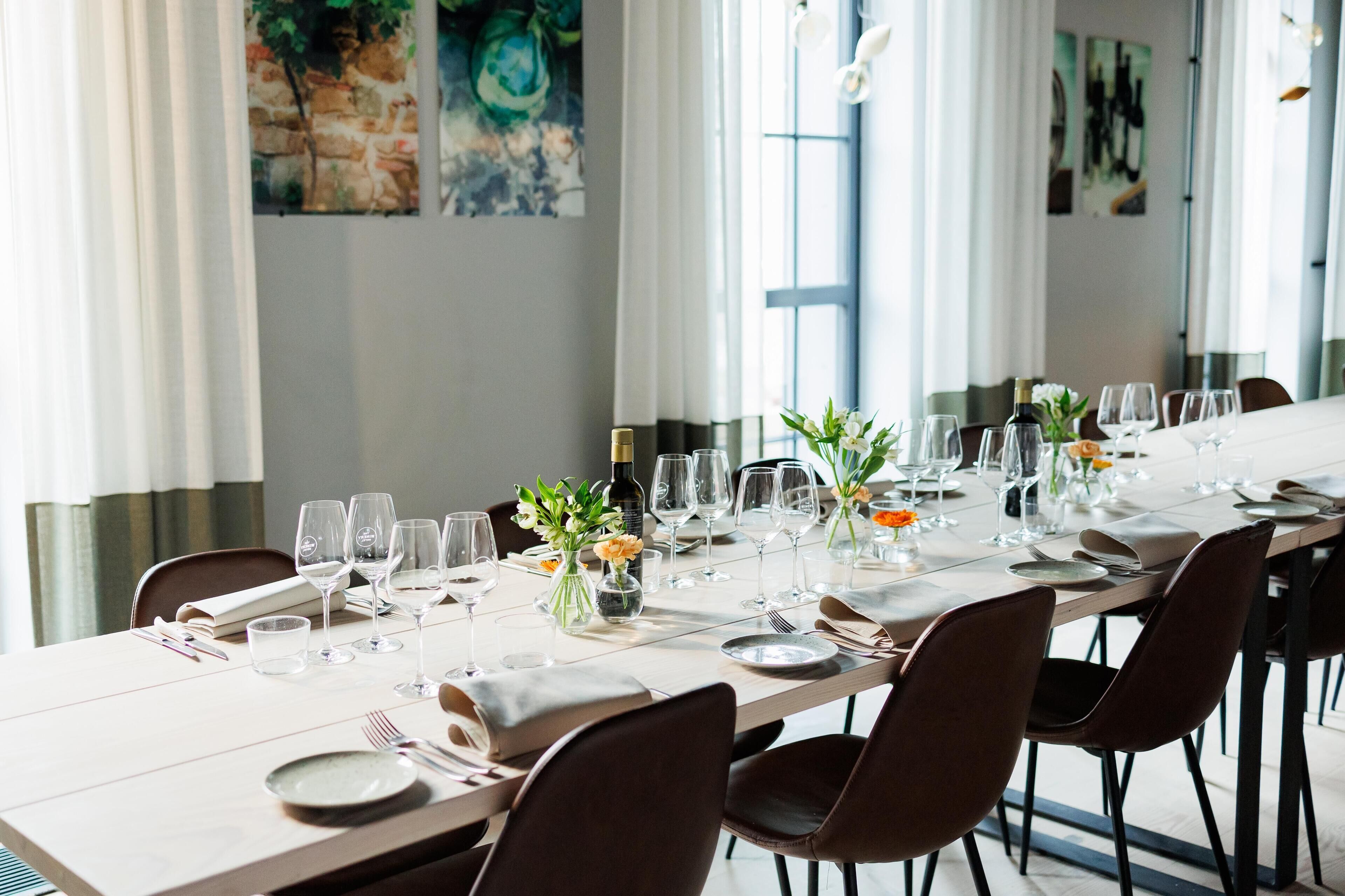 Una larga mesa de comedor dispuesta con copas de vino, platos y jarrones con flores, en una habitación bien iluminada con ventanas grandes y obras de arte en las paredes.