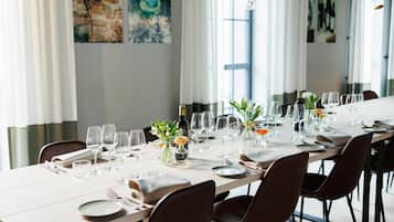 Una larga mesa de comedor dispuesta con copas de vino, platos y jarrones con flores, en una habitación bien iluminada con ventanas grandes y obras de arte en las paredes.