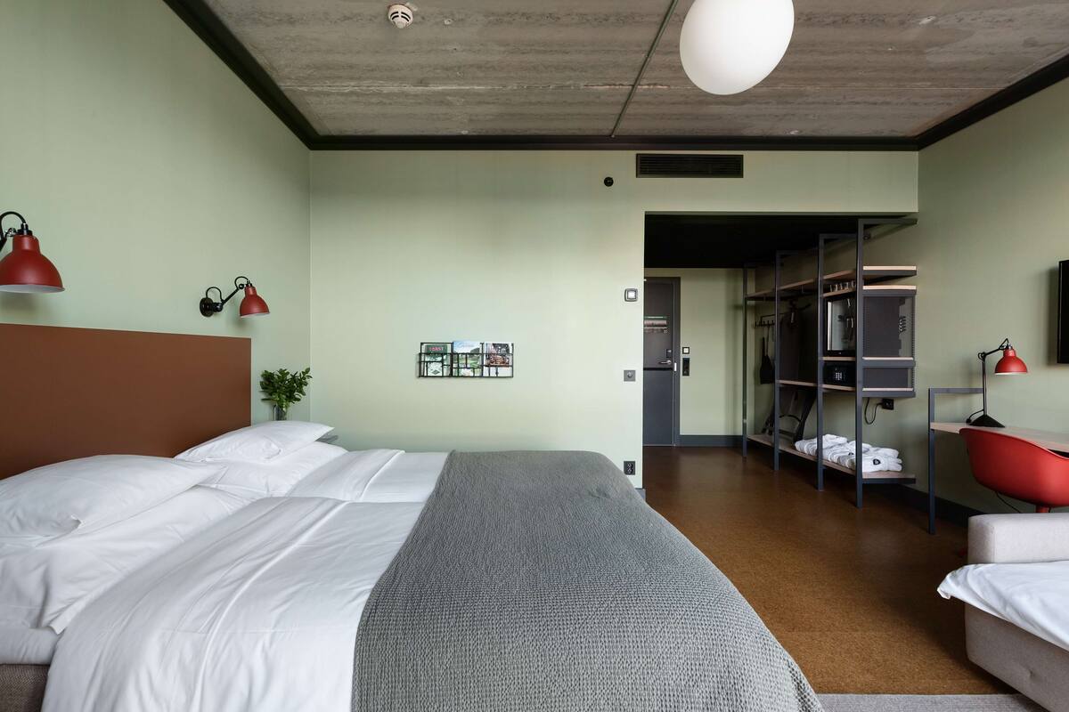 Ein Hotelzimmer mit Bett, Nachttischen, einer roten Lampe, einer grauen Decke, einer Pflanze und einem Kleiderschrank.