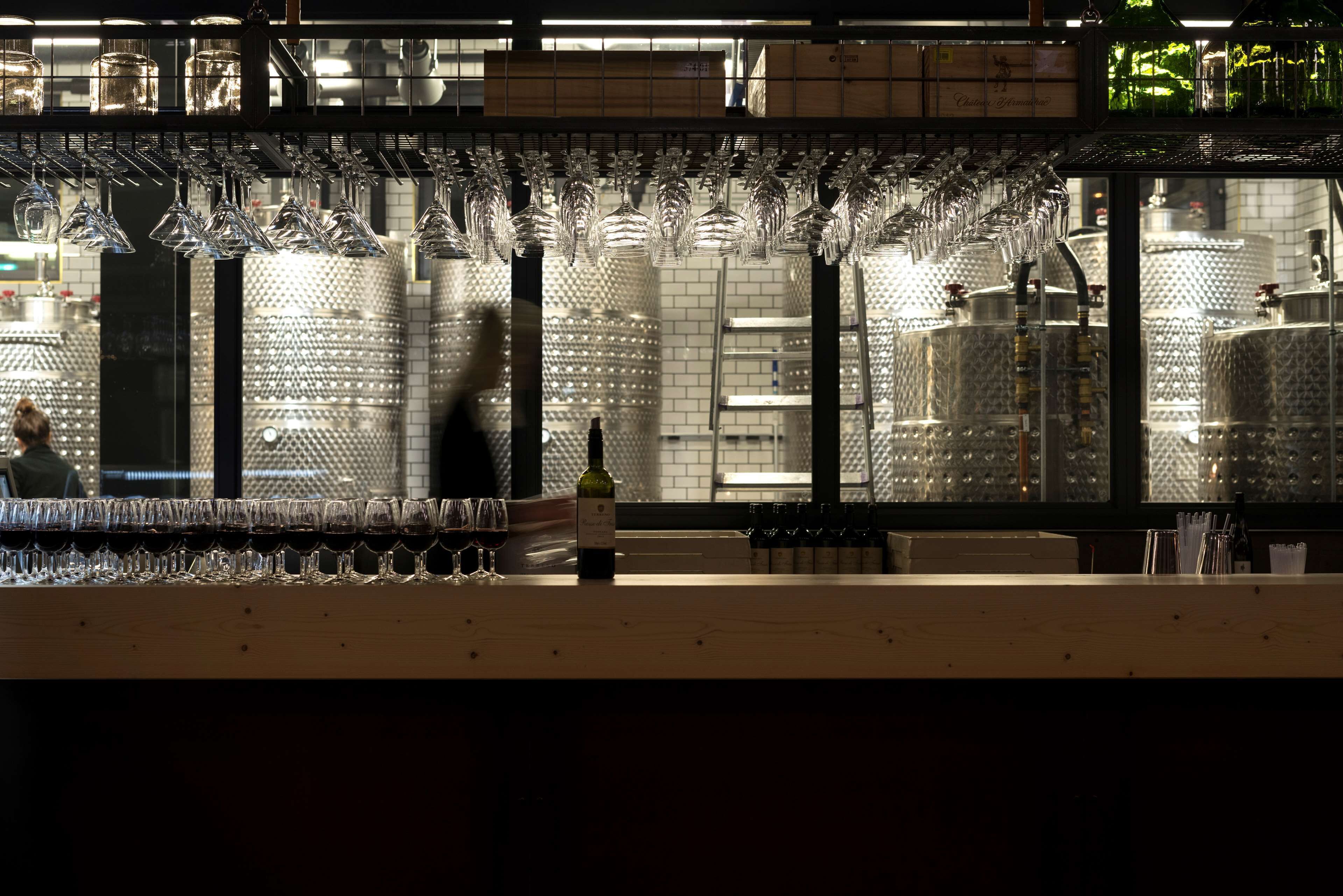 A bar area with multiple hanging wine glasses, a bottle of wine, and a person in the background.