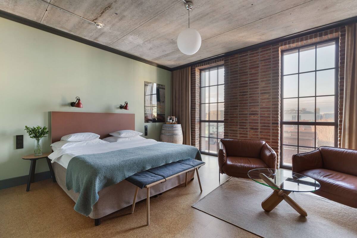 Ein modernes Hotelzimmer mit einem großen Bett, einem Glastisch, Ledersesseln und Blick auf die Stadt durch große Fenster.