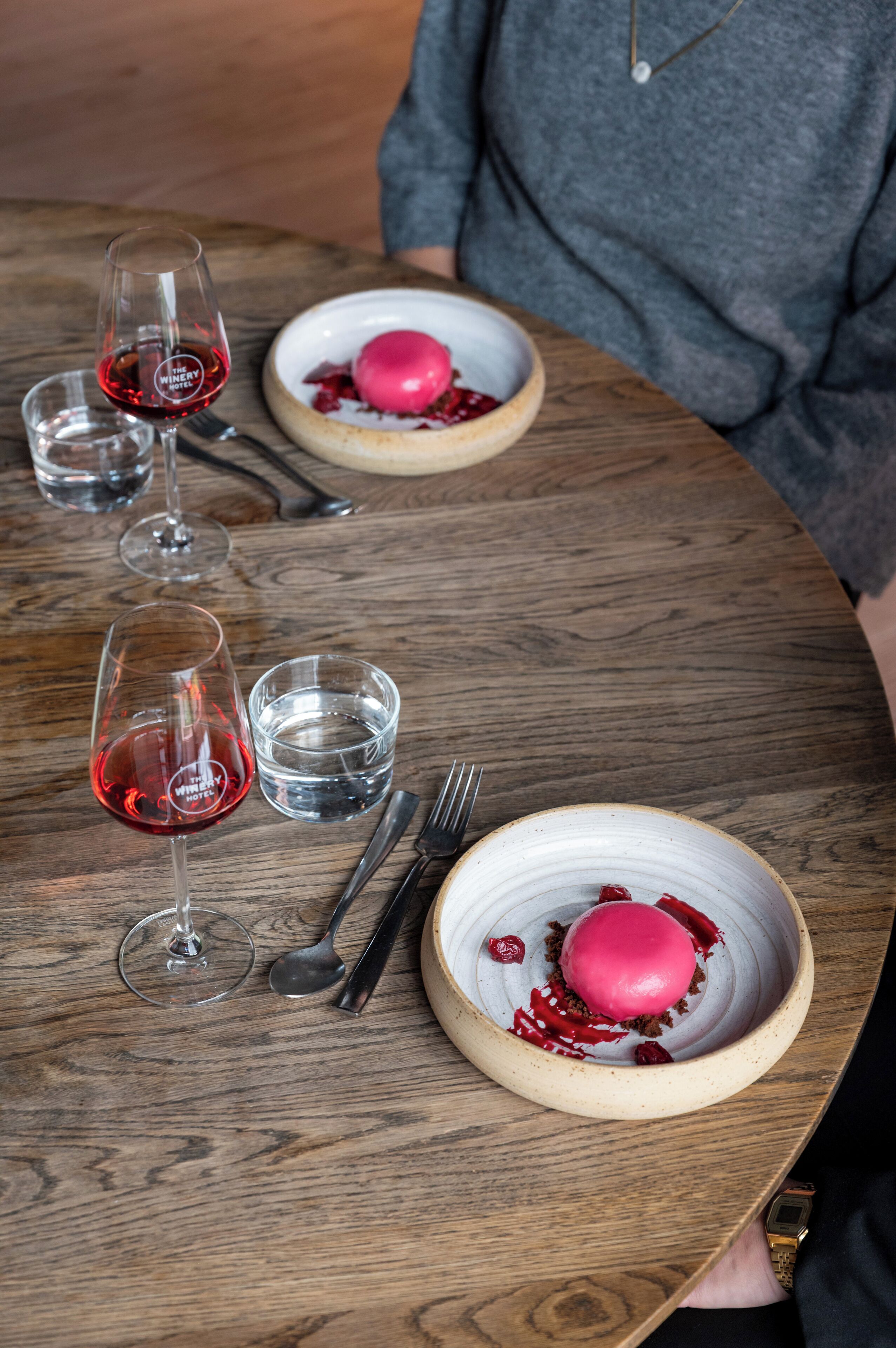 A wooden table with a dessert, wine glasses, and cutlery.