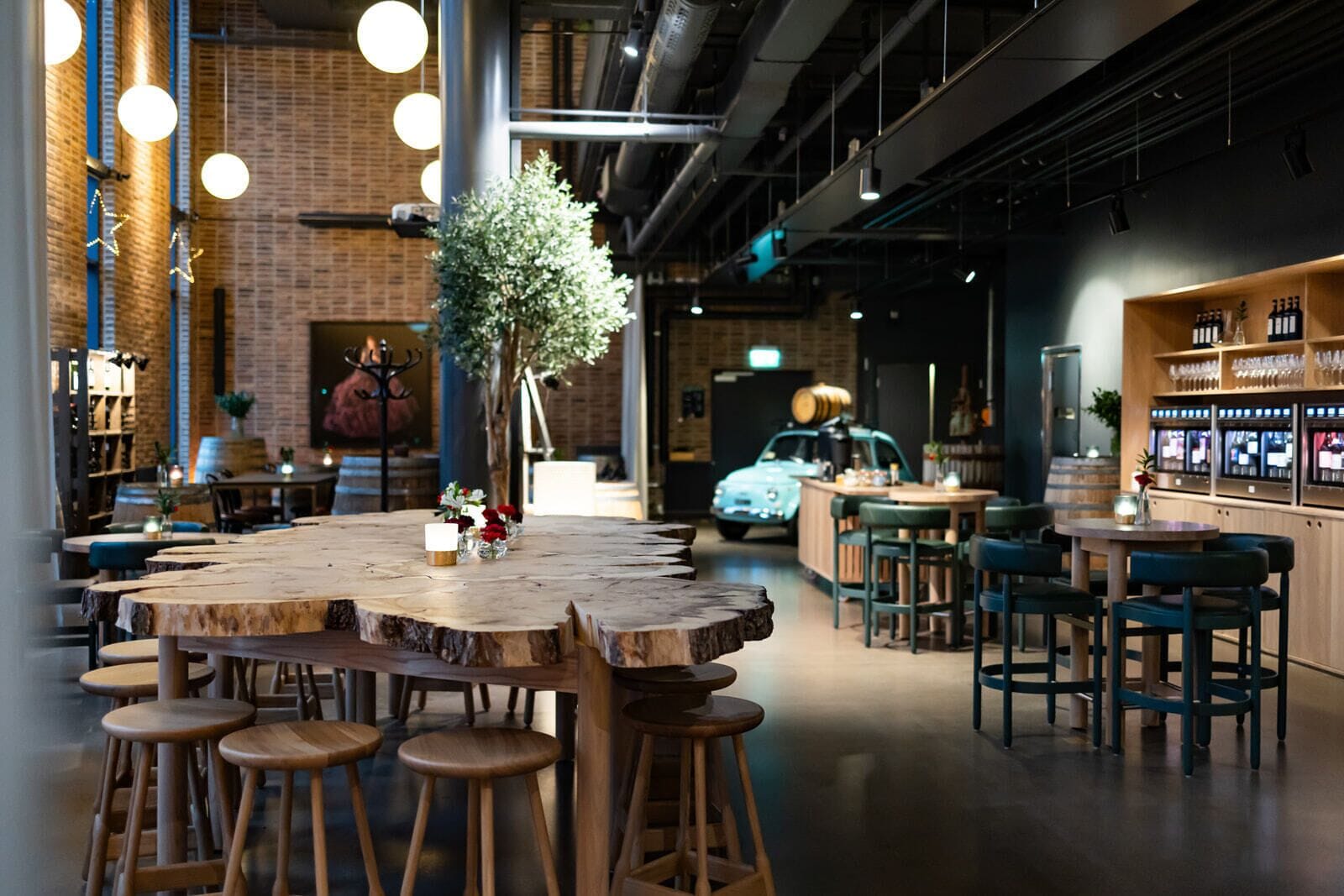 A modern bar area with a central wooden table surrounded by stools, a bar counter with glasses and bottles, and a vintage car in the background.