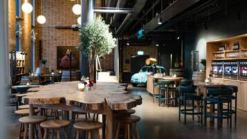A modern bar area with a central wooden table surrounded by stools, a bar counter with glasses and bottles, and a vintage car in the background.