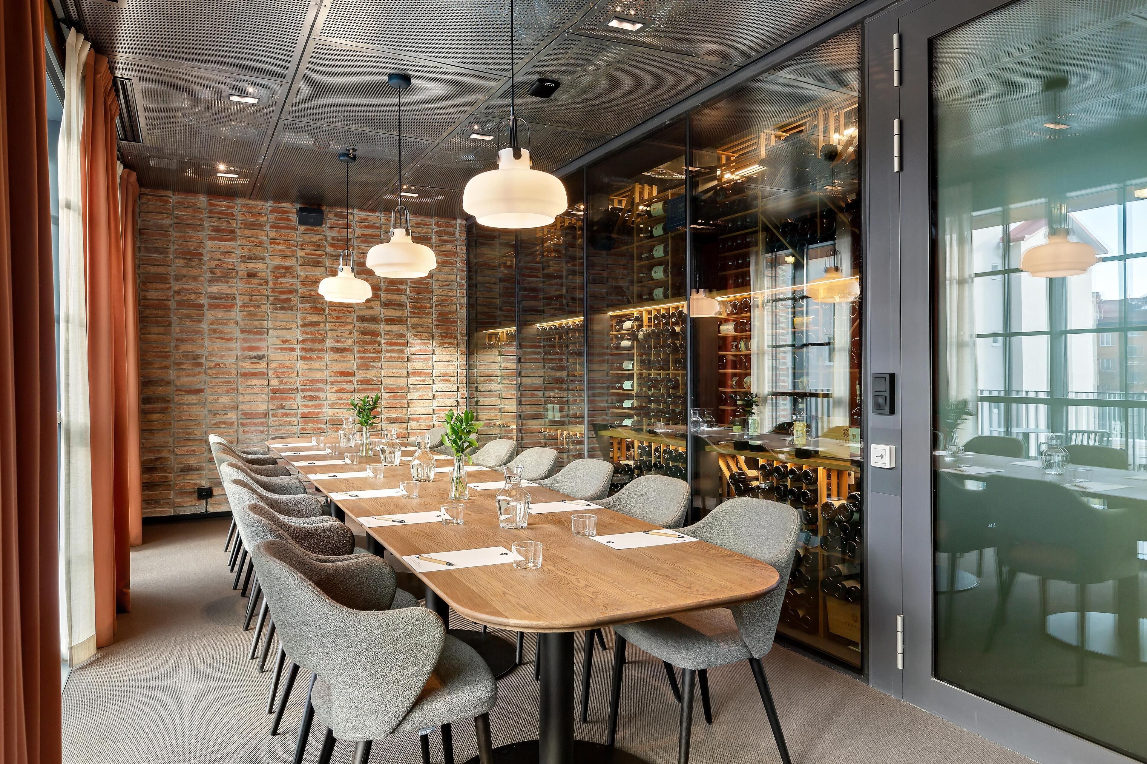 A conference room with a long wooden table, grey upholstered chairs, hanging pendant lights, and a brick wall with a wine rack visible through a glass partition.
