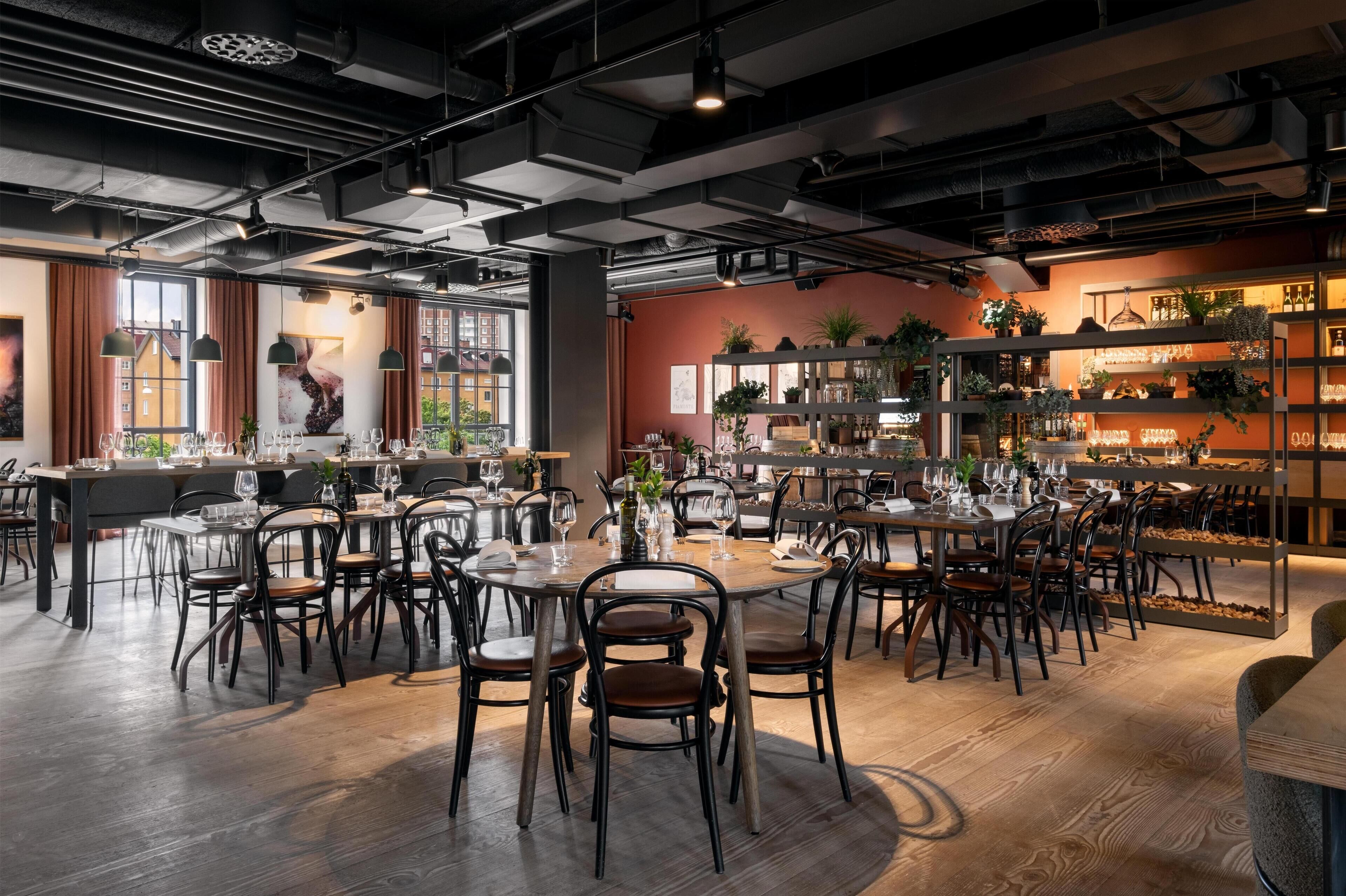 A modern restaurant with multiple dining tables and chairs, a bar area with shelves of bottles, and large windows allowing natural light.