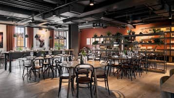 A modern restaurant with multiple dining tables and chairs, a bar area with shelves of bottles, and large windows allowing natural light.