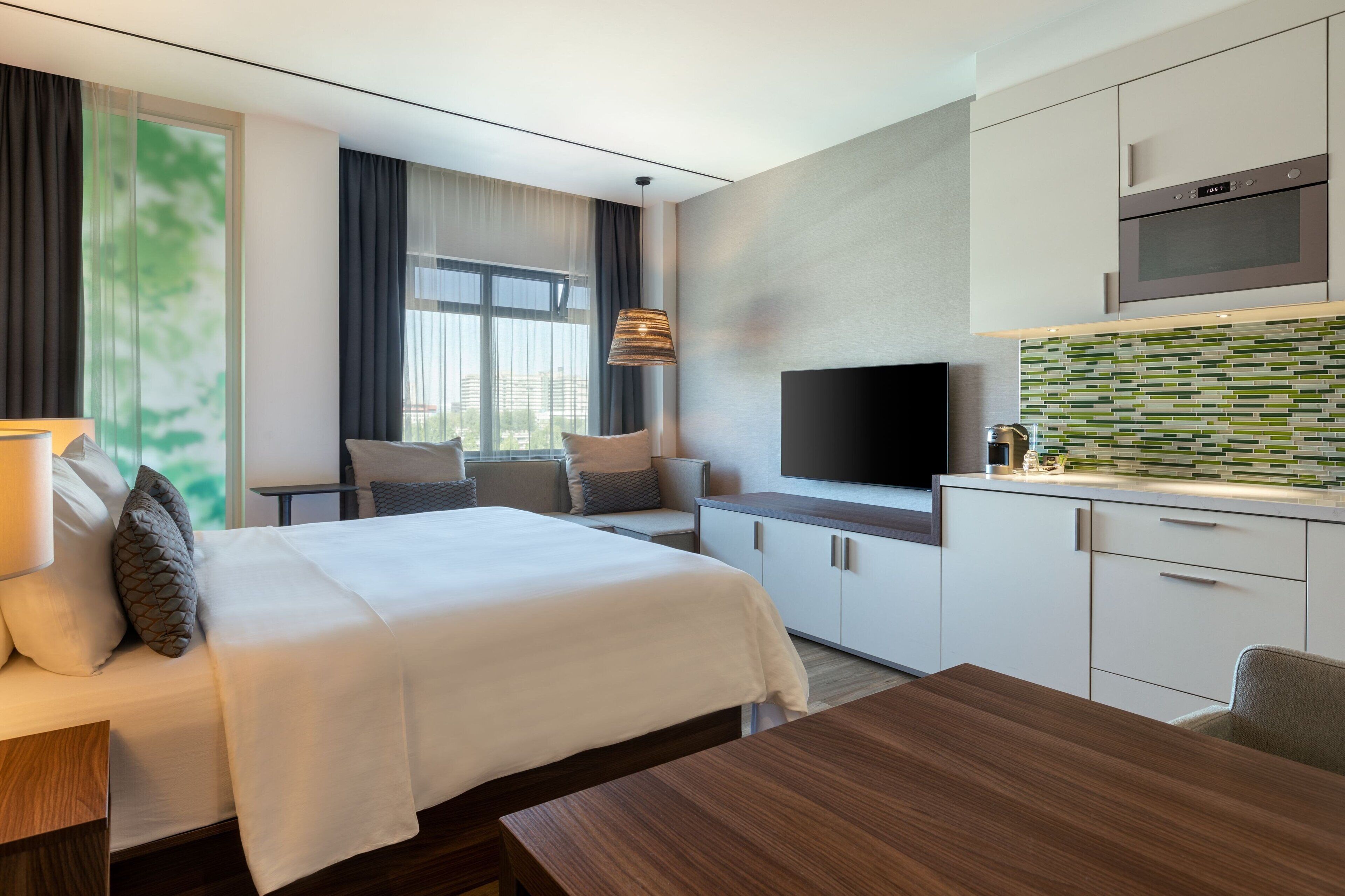 A modern hotel room with a bed, a desk, a TV, and a kitchenette.