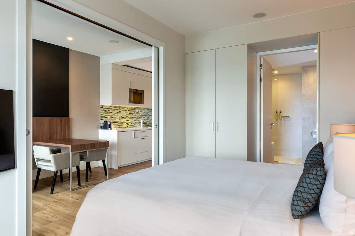 A modern hotel room with a large bed, a desk, and a kitchenette in the background.