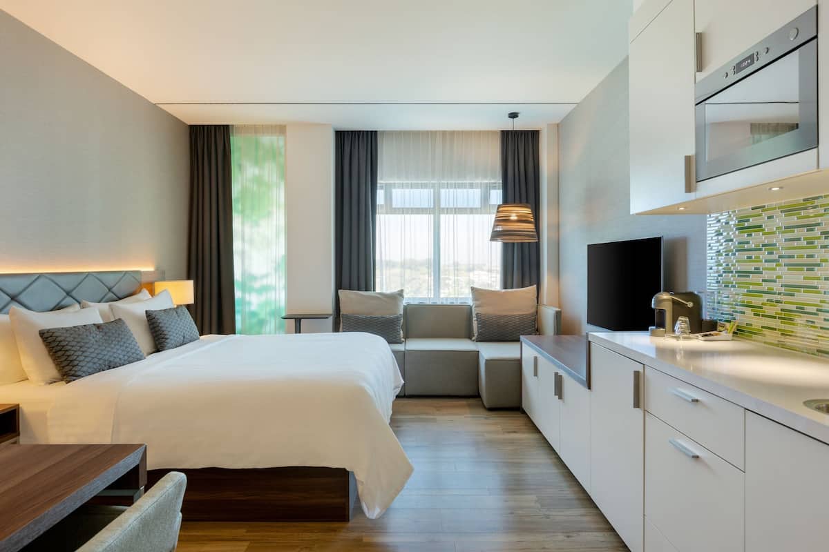A modern hotel room with a large bed, a seating area, a kitchenette, and a TV.