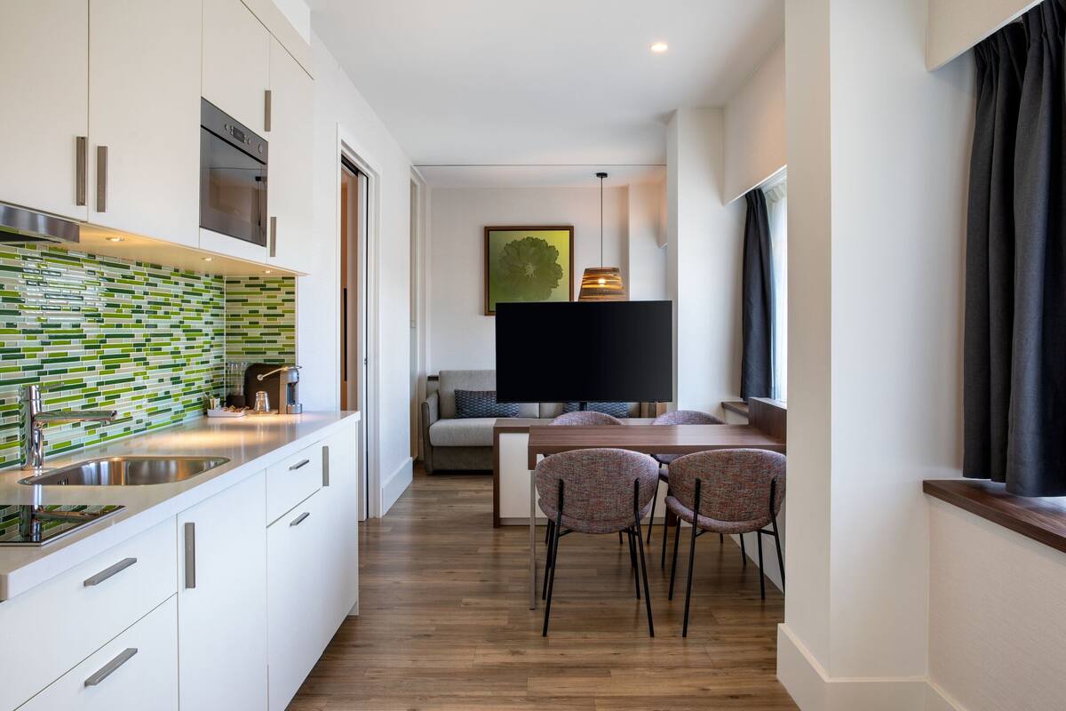 A modern kitchen with white cabinets, a stainless steel sink, and a green and white backsplash. There is a dining area with a table and chairs, and a flat-screen TV mounted on the wall.