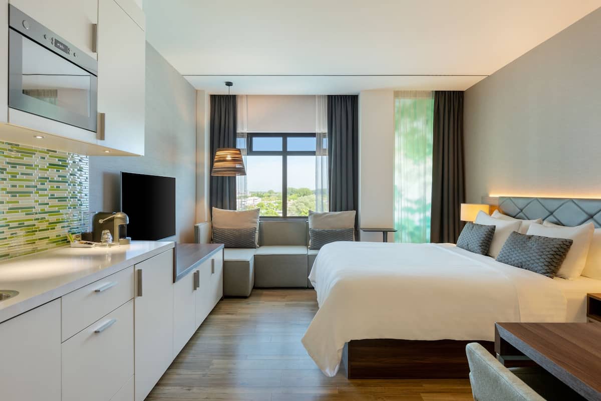 A modern hotel room with a bed, seating area, kitchenette, and a large window providing a view.