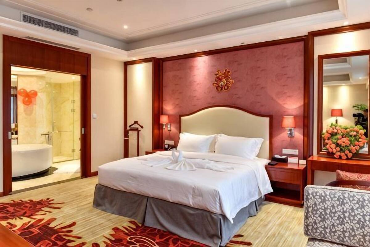 A hotel room with a large bed, a sitting area, a bathroom with a bathtub, and a mirror with a flower arrangement.