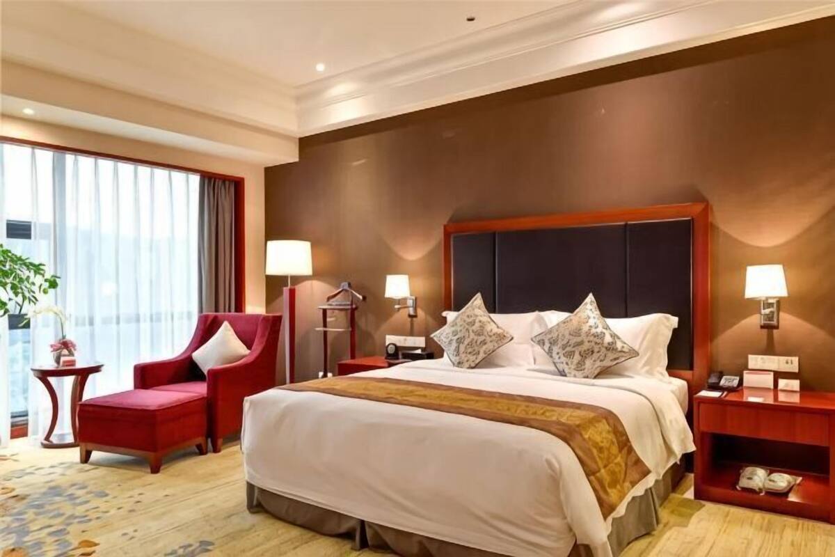 A hotel room with a large bed, two bedside tables, a red armchair, a nightstand, and a view of the city.
