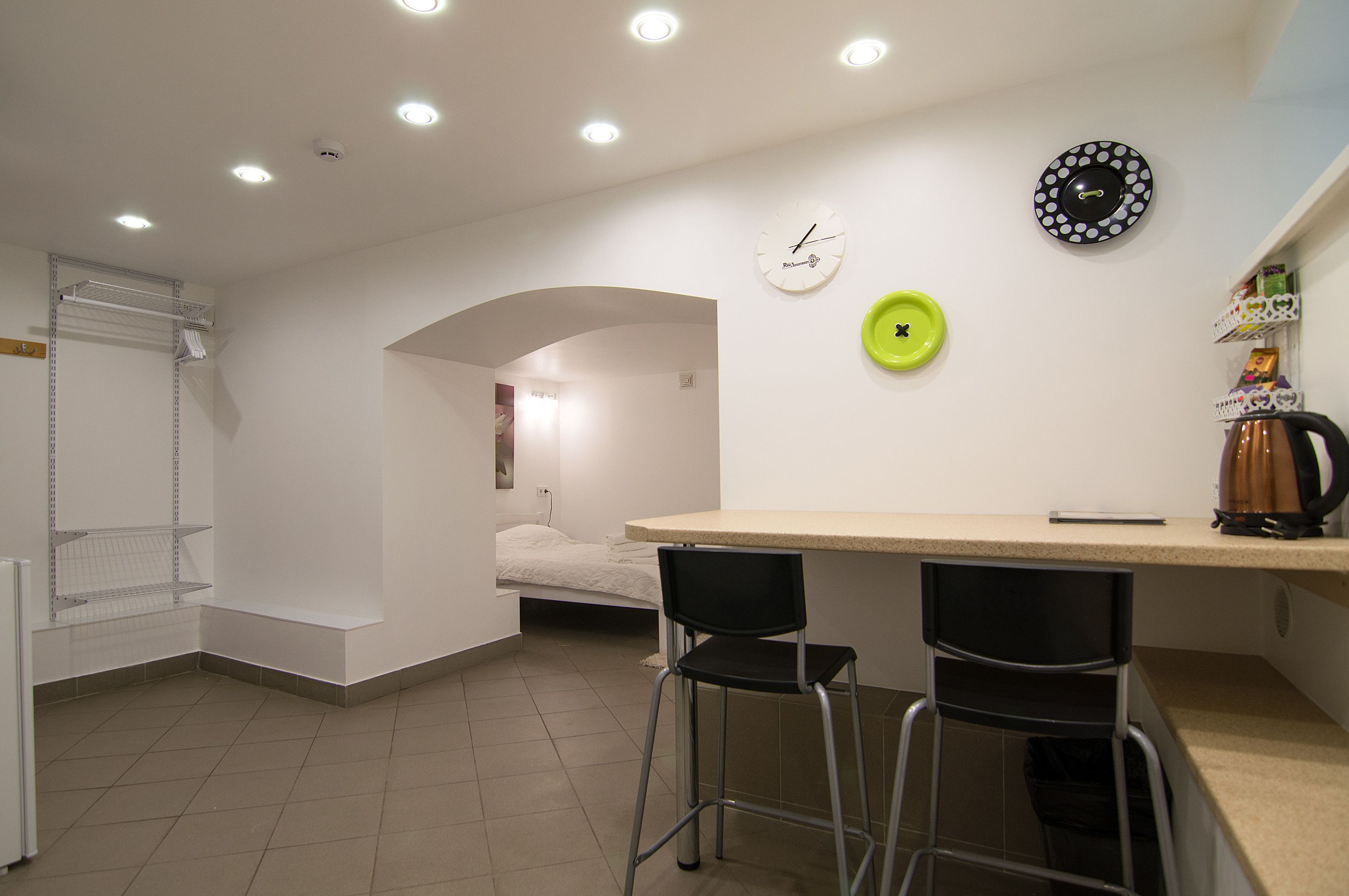 A compact kitchen with a small dining area, a bed in the background, and a clock on the wall.